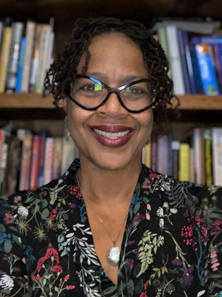 Headshot of Sonya Grier, sitting in front of a bookcase, smiling and wearing a black floral shirt